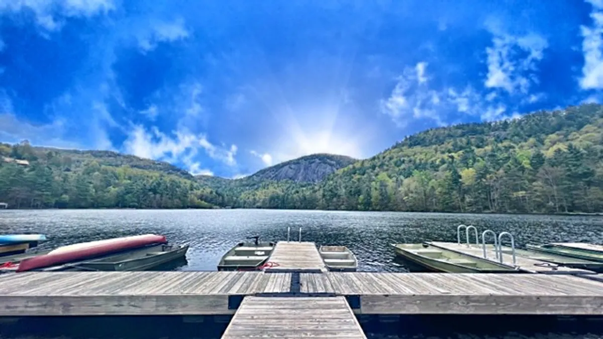 A peaceful lakeside dock stretches into the water, surrounded by boats and with majestic mountains rising in the background. The sky above is clear, suggesting a serene day ideal for outdoor activities. The natural scenery is lush and inviting, hinting at the abundance of trails and exploration routes nearby.