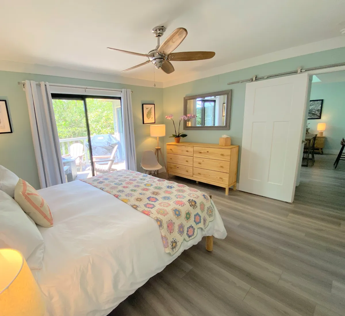 A serene bedroom featuring a bed with a patterned blanket, a wooden dresser, and a large window offering a tranquil view of the outdoors. Soft natural light fills the space, creating a cozy and inviting atmosphere ideal for rest and relaxation.