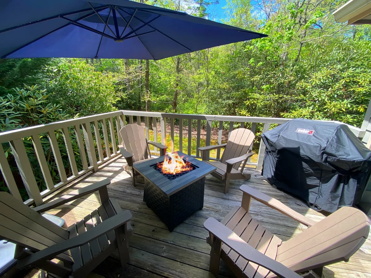 The image shows a cozy outdoor setting at The Sapphire Retreat, with Adirondack chairs arranged around a glowing fire pit, an outdoor umbrella and grill nearby, all framed by lush mountain greenery. The scene exudes warmth, inviting guests to relax, share stories by the fire, and enjoy sunset views from the deck.