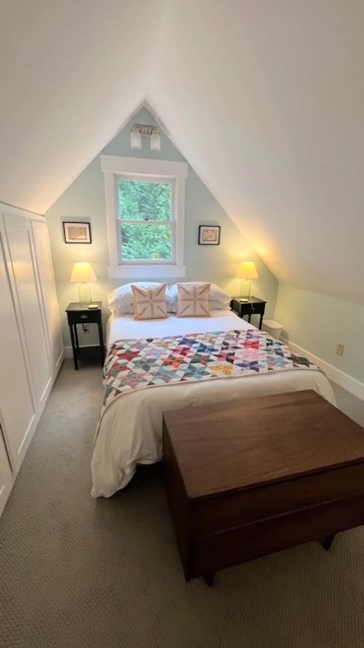 An inviting bedroom with a bed dressed in a colorful quilt, nightstands with lamps on each side, and a window that lets in soft, natural light. The decor is simple yet warm, evoking a sense of comfort and private retreat.