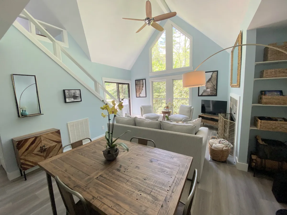 A spacious, welcoming open-plan living and dining area with high ceilings and large windows that let in abundant natural light. The scene includes a wooden dining table perfect for shared meals, a comfortable seating area for relaxing, and decorative art pieces that contribute to a warm, inviting atmosphere.