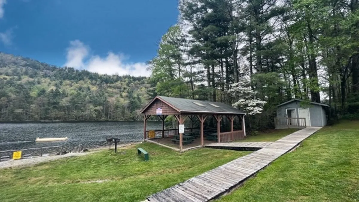 A peaceful lake scene features a charming gazebo, a curving pathway, and tall surrounding trees. The calm water reflects the soft light, inviting activities like fishing, boating, or simply relaxing by the shore.