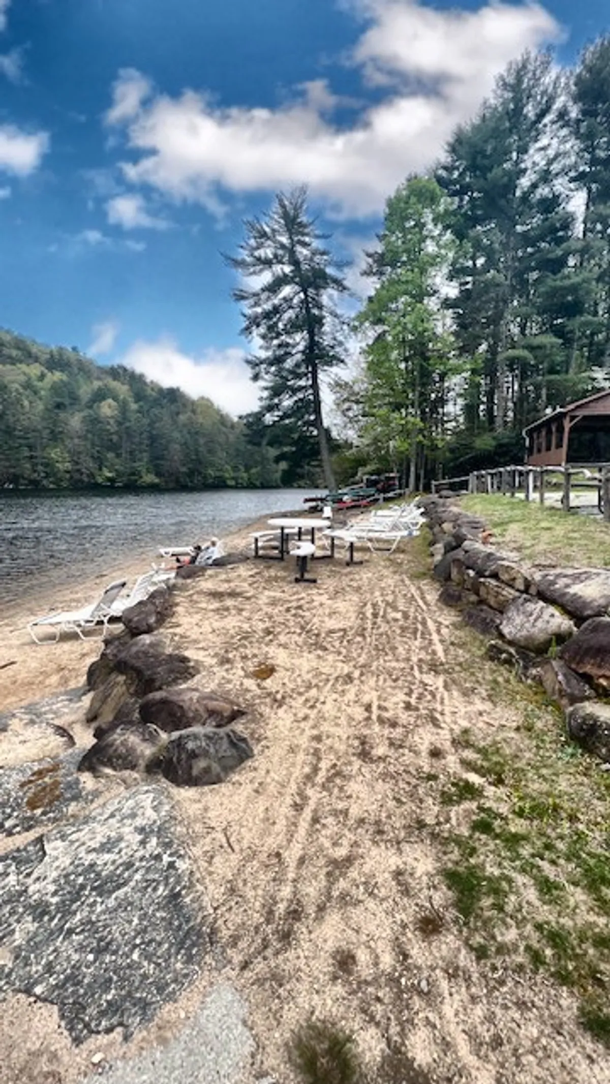 This image captures a beautiful lake surrounded by trees, with a sandy beach area, lounge chairs, and picnic tables in the foreground. The scene suggests opportunities for swimming, boating, sunbathing, and enjoying outdoor meals. The tranquil waters and natural setting embody the recreational spirit of the Sapphire Rec Center mentioned in the section.