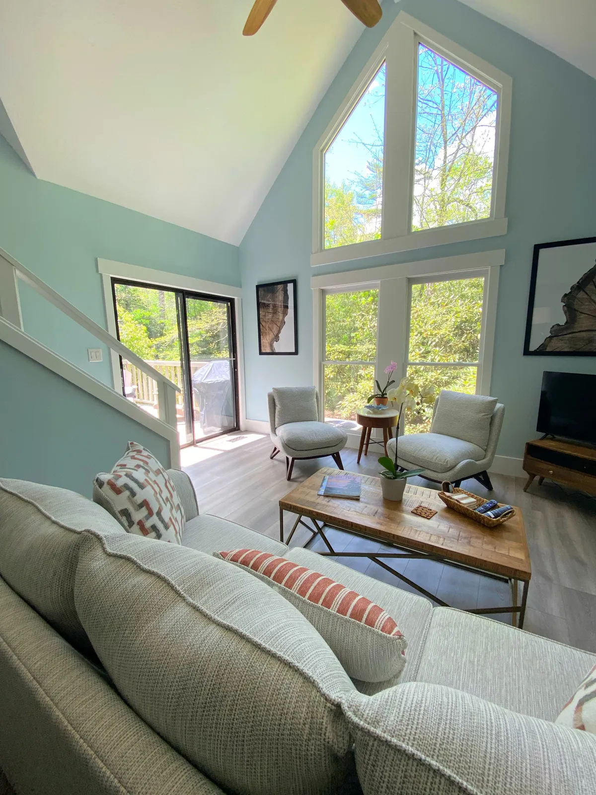 This image displays a cozy living space featuring large windows that invite natural light, comfortable seating, a wooden coffee table, and lush decorative plants. The view through the windows hints at the surrounding wooded setting, connecting the interior to the outdoors.