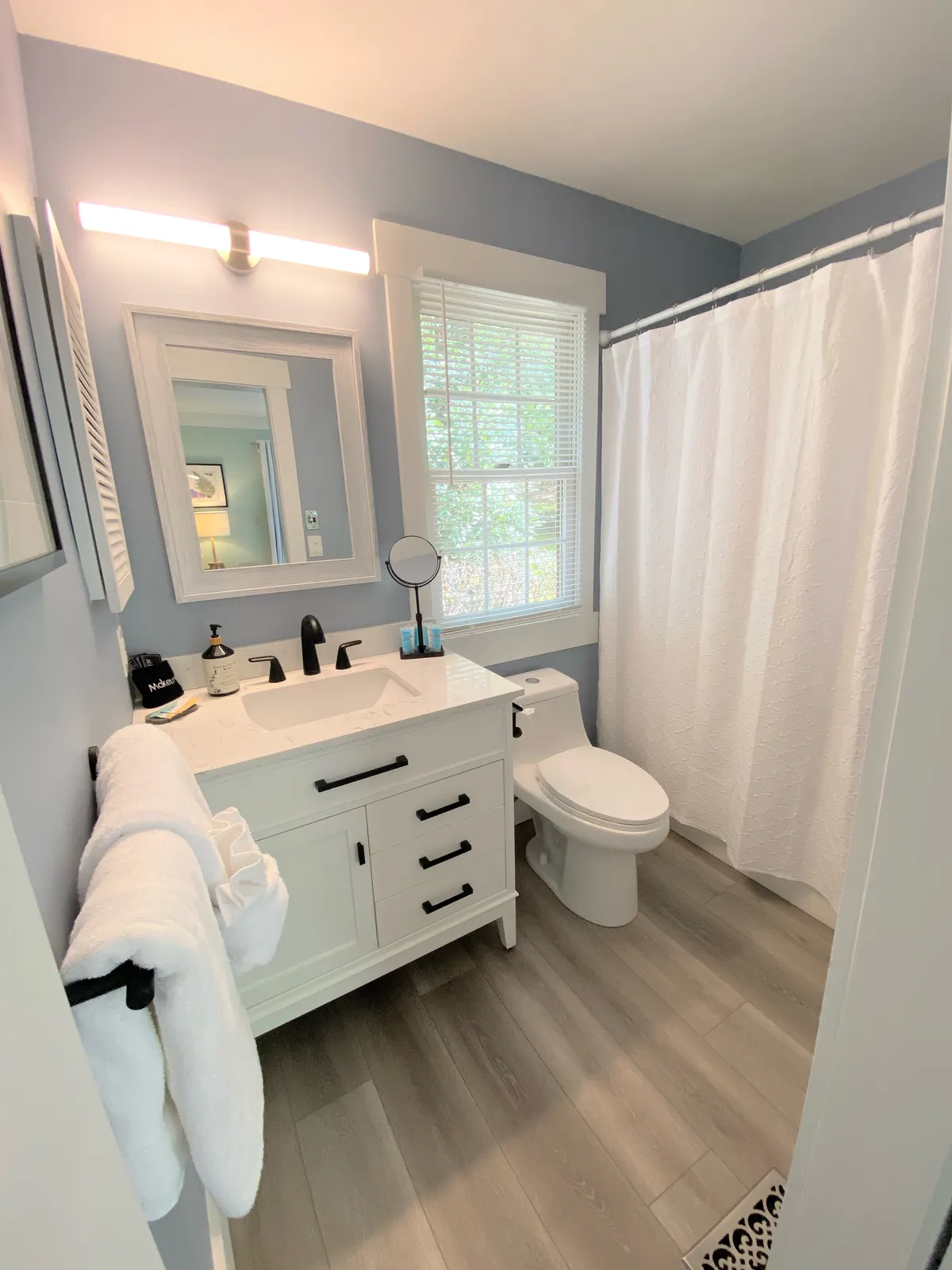 A bright, calming en-suite bathroom complete with a vanity sink, mirror, window, shower curtain, and toilet. The layout is both functional and inviting, providing a peaceful space for morning routines or evening wind-downs.