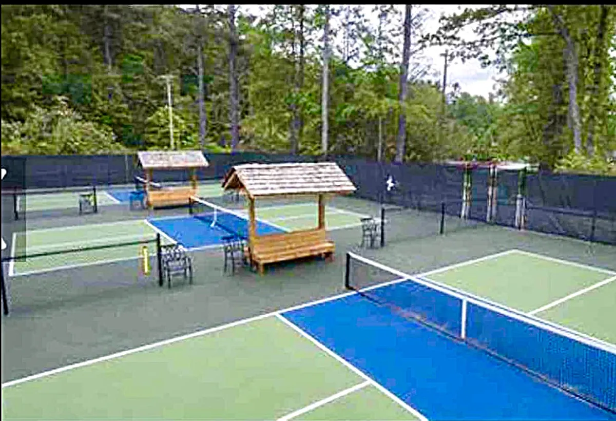 Well-maintained tennis courts are framed by tall leafy trees, with sheltered seating areas nearby. The setting is open and inviting, surrounded by greenery and dappled sunlight, evoking a sense of recreation and connection to the outdoors.