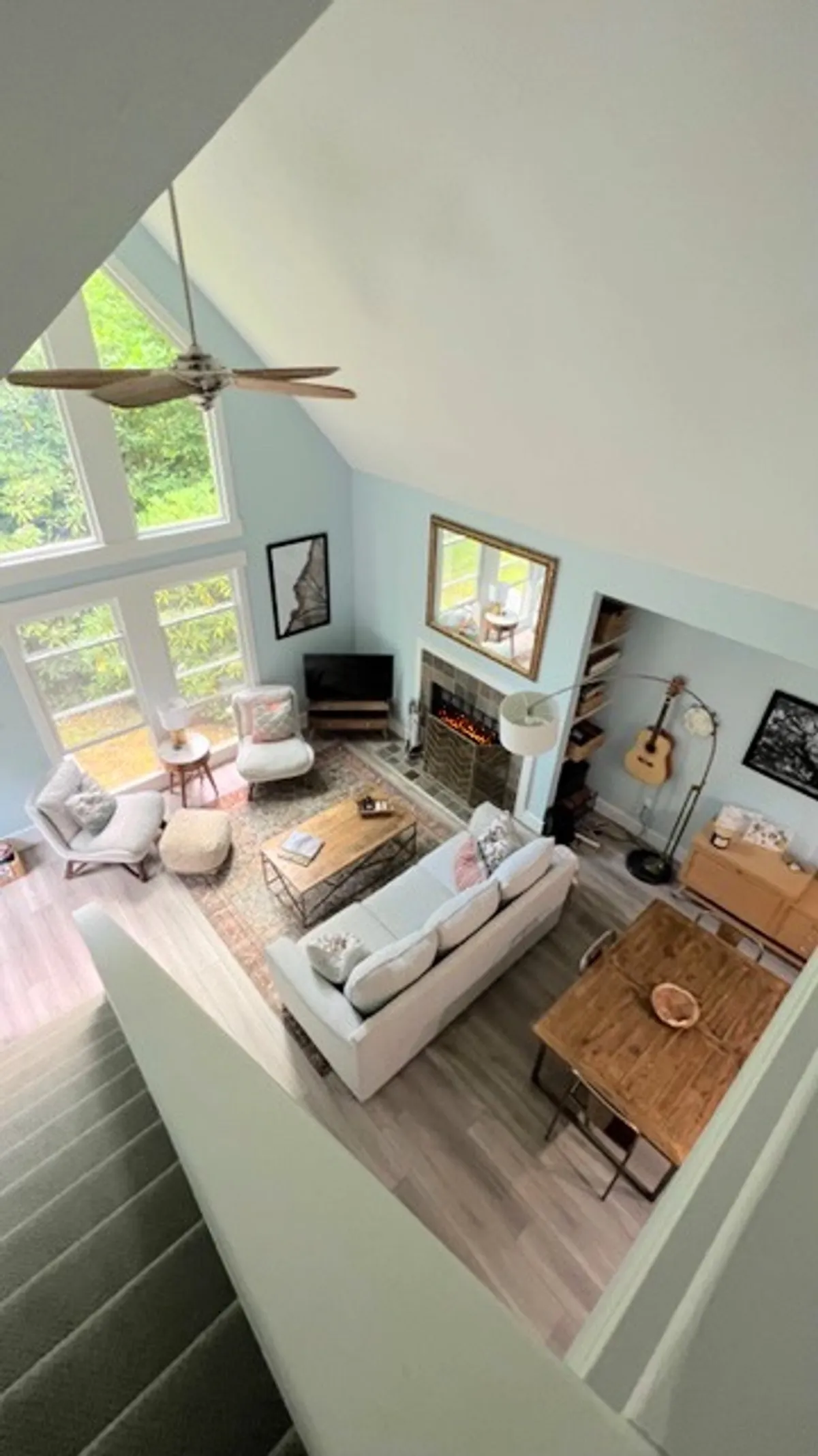 A spacious, light-filled central living area featuring a cozy sofa and coffee table near a fireplace. A wooden dining table sits in the background, with a guitar resting beside it. Sunlight streams through large windows, and tasteful decor enhances the sense of comfort and cleanliness.