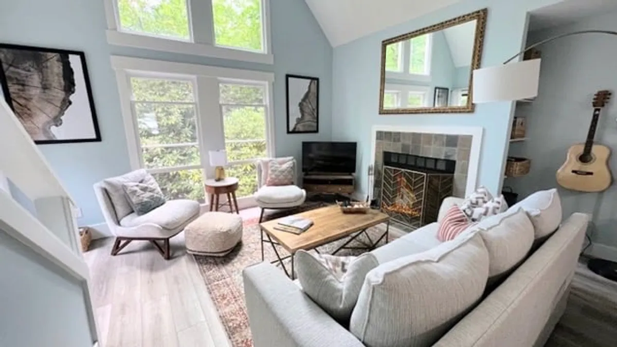 A bright, inviting living room with a comfortable sofa and coffee table arranged around a fireplace. A guitar leans nearby, and large windows fill the space with natural light, enhancing the cozy, updated atmosphere.